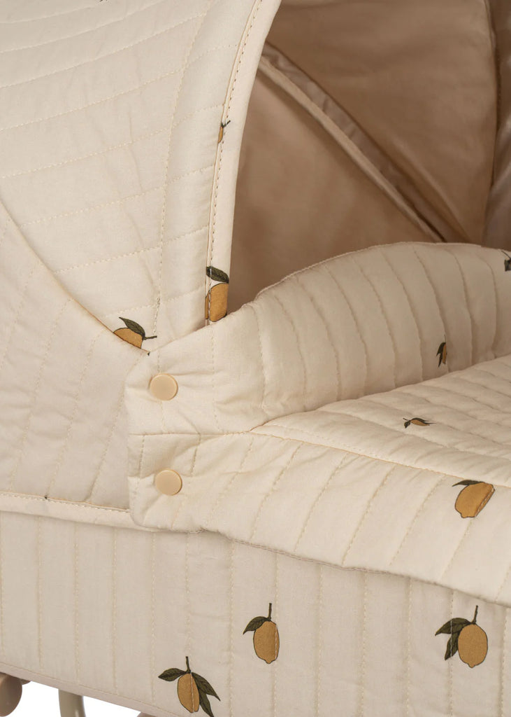 A close-up of the Konges Sloejd Mini Doll Pram in a beige shade showcases its quilted fabric adorned with a lemon pattern. This bassinet, reminiscent of vintage doll prams, features two buttons on the side, highlighting its cozy and decorative design. (Ships in approximately one week.)