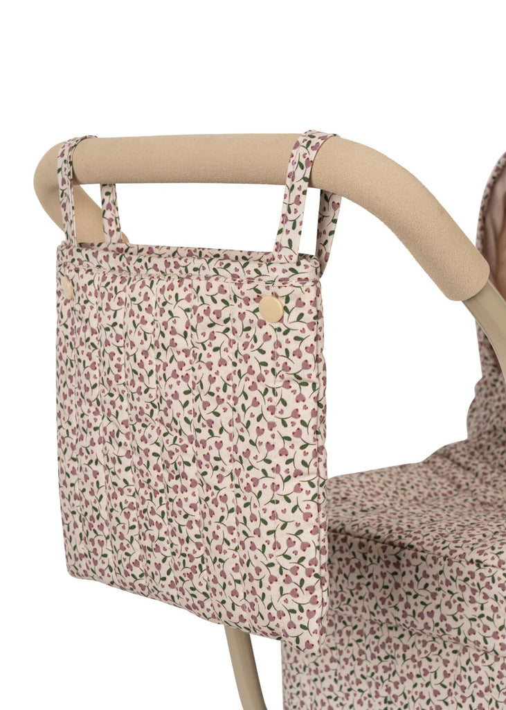 The Konges Sloejd Mini Doll Pram - Milk Tank features a close-up of the stroller handle with a floral-patterned fabric bag attached, highlighting its quilted fabric design. The vibrant floral print on a light background complements the cushioned storage basket, which includes button detail that matches the stroller seat fabric.