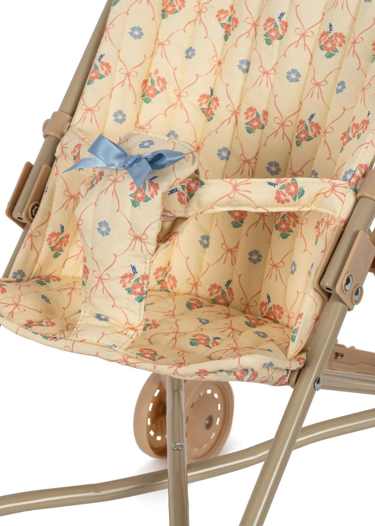 A detailed view of the Konges Sloejd Doll Stroller - Fleur Decor reveals its empty design, characterized by a beige metal frame and double wheels. The stroller features an elegant floral pattern adorned with a blue bow and includes removable fabric for effortless cleaning.