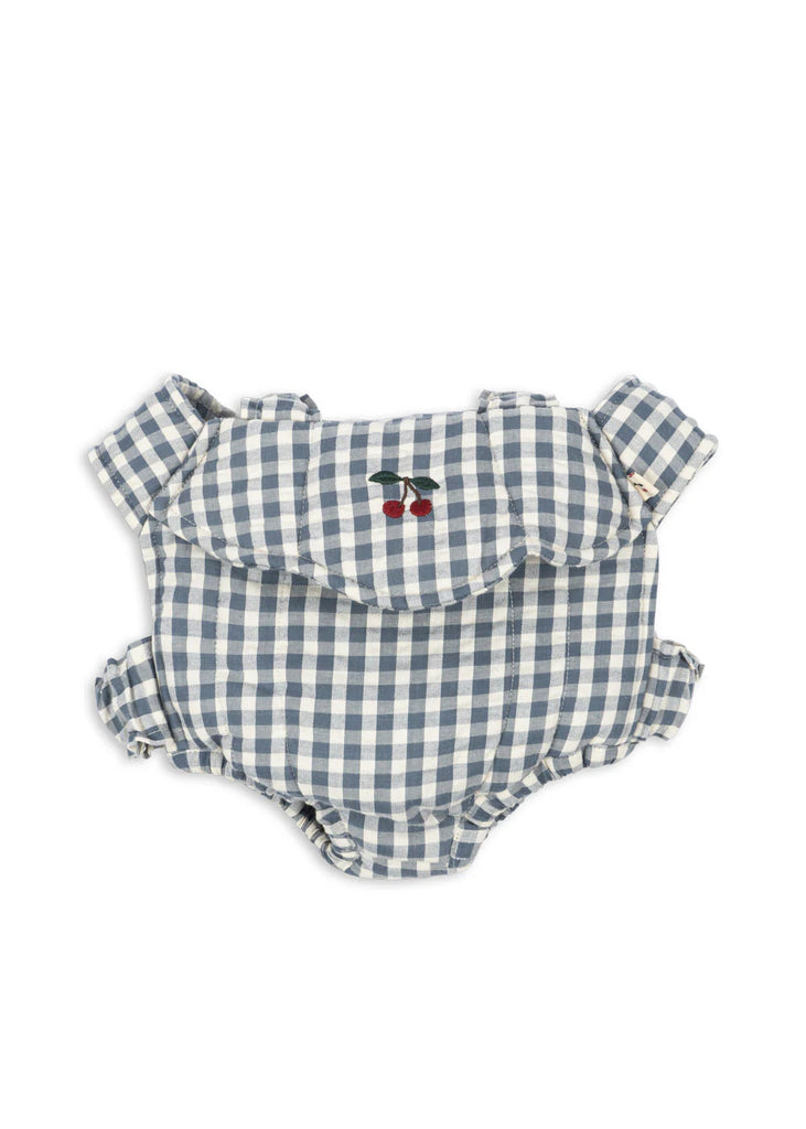 A baby romper in blue and white gingham with ruffled sleeves and an embroidered cherry on the chest, shown next to the Konges Sloejd Doll Carrier - Blue Checkers, all set against a plain white background.