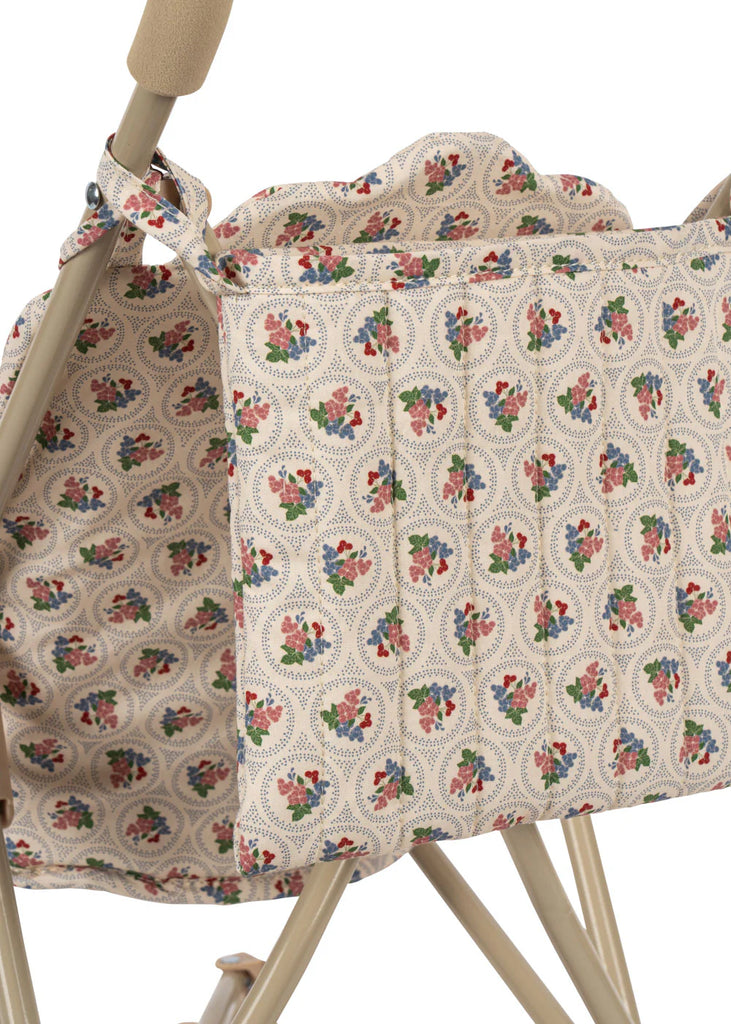 The Konges Sloejd Doll Stroller - Verona Blues features a close-up of its red, blue, and green floral-print carriage bag on beige fabric, draped over the handle of a beige metal doll stroller with double wheels.