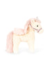 The Konges Sloejd Starlight Unicorn Stuffed Animal features cream-colored fur, a pink mane and tail, pink wings, a silver horn, and a floral-patterned harness, and is shown standing upright on all four legs.