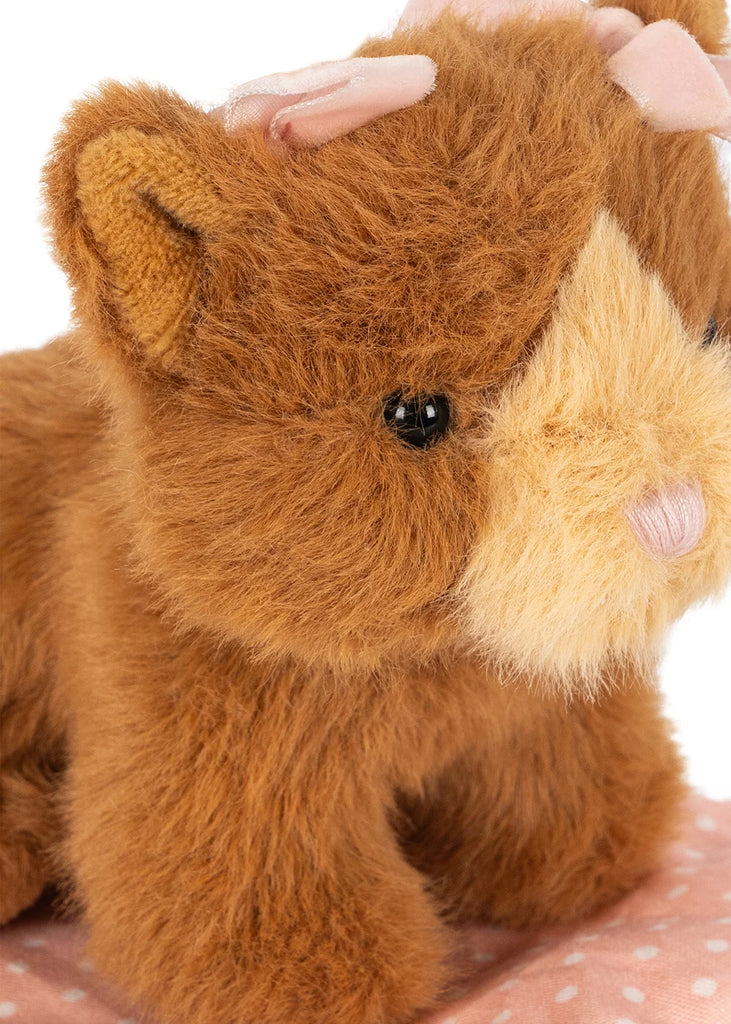 A close-up of the Konges Sloejd Take Care of Me Animals - Cat, featuring a brown and beige plush kitten with a pink bow on its head, sitting on a pink fabric with white polka dots. This adorable mini toy is perfect for any collection from the ‘Take Care of Me’ series.