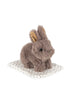A petite, plush gray bunny with tan inner ears from the Konges Sloejd Take Care of Me Animals collection sits atop a white cloth dotted with tiny black spots, embodying the charm of a perfect mini toy animal and softly inviting you to “take care of me.”.