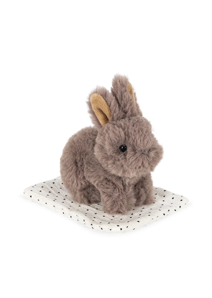 A petite, plush gray bunny with tan inner ears from the Konges Sloejd Take Care of Me Animals collection sits atop a white cloth dotted with tiny black spots, embodying the charm of a perfect mini toy animal and softly inviting you to “take care of me.”.