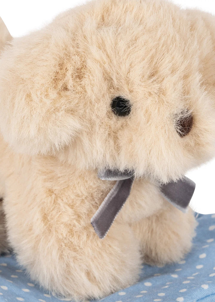 A Konges Sloejd Take Care of Me Animals - Dog plush, featuring soft beige fur, black eyes, a brown nose, and a gray bow around its neck, sits on light blue fabric with white polka dots, gazing upward in an endearing pose.