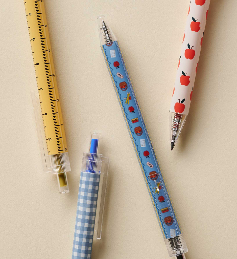 The Schoolhouse Gel Pen Set of 4 includes four retractable gel pens in unique designs—yellow with ruler markings, blue checkered, blue with school-themed icons, and white with red apples—arranged on a light beige surface. Each pen is equipped with a 0.5 mm rollerball tip for smooth writing and features vibrant ink that brings a pop of color to your notes.