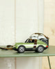 A Playforever Car LUFT HOPPER Green with black wheels rests on a green table. A vintage camera appears in the background against a light wall, highlighting the collectible charm of this stylish toy car display.