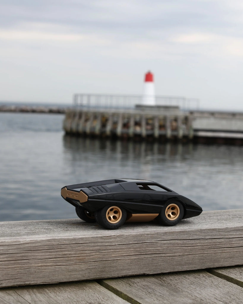 The Playforeer UFO Cannone Car in Black, a small toy with gold wheels inspired by 70s concept cars, rests on a wooden surface by the water, with a lighthouse and dock visible in the cloudy background.