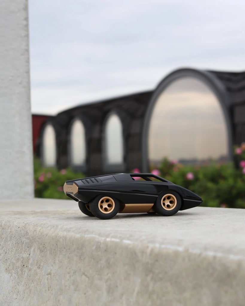 The Playforeer UFO Cannone Car in Black, a small toy inspired by 70's concept cars, rests on a stone ledge outdoors with blurred arched glass buildings and greenery in the background beneath a cloudy sky.