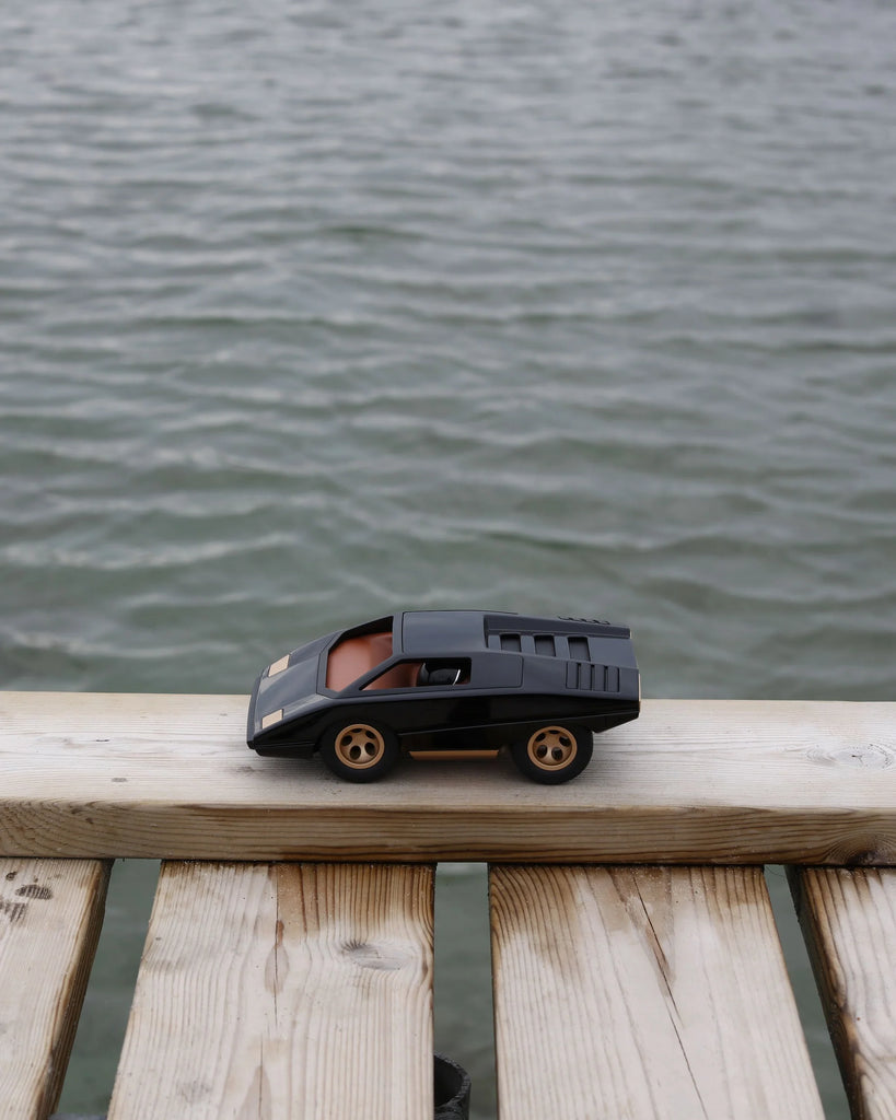 The Playforeer UFO Cannone Car in Black, inspired by 70's concept cars, sits on a wooden dock above the water with rippling waves in the background.