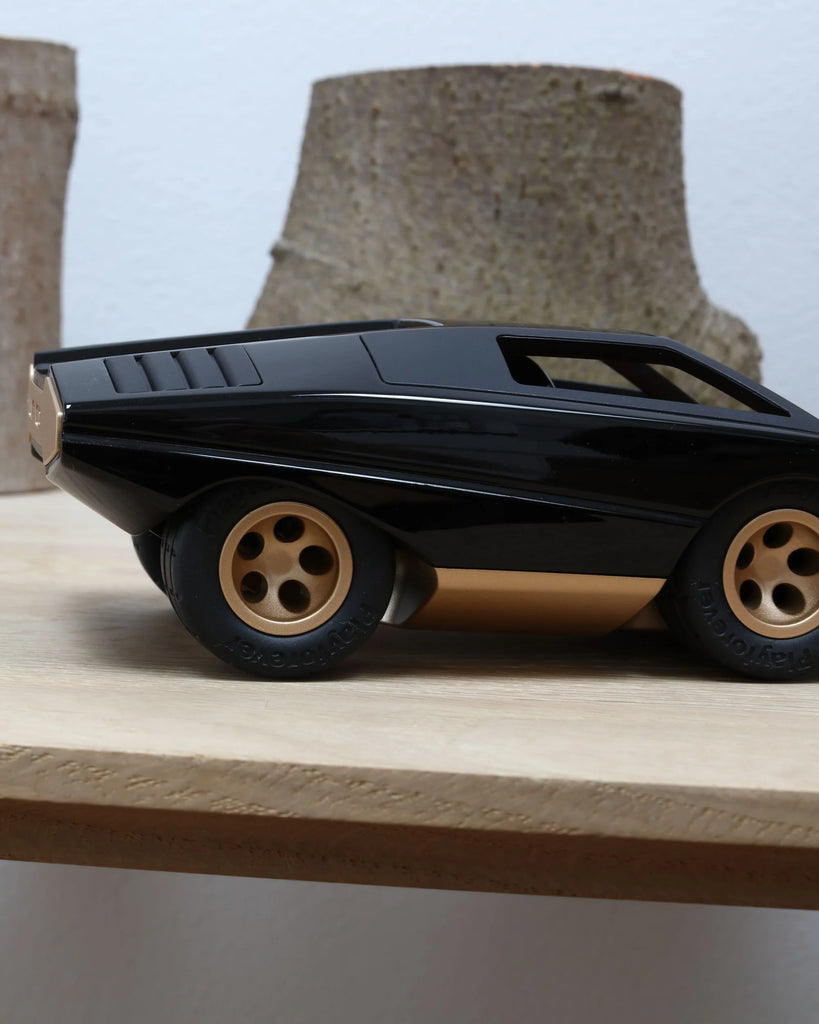 The Playforeer UFO Cannone Car in Black, featuring bold 70's-inspired lines and large wheels, is displayed on a wooden surface against a backdrop of blurred tree trunk segments.