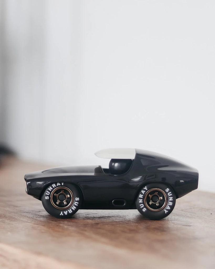 A Playforever Leadbelly Skeeter Car in Black, featuring a white roof and "SUNRAY" on its tires, sits on a wooden surface against a plain, light background.