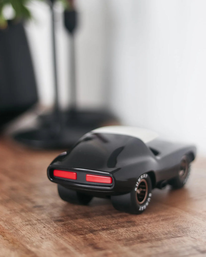 The Playforever Leadbelly Skeeter Car in Black, a small toy car with red tail lights, sits on a wooden surface. A blurred background shows a lamp and greenery.