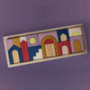 The Grimm's Panorama Building Set features vibrant geometric wooden blocks in blue, red, yellow, and natural tones, forming abstract houses and arches on a wooden tray—creating a lively scene against a purple background.