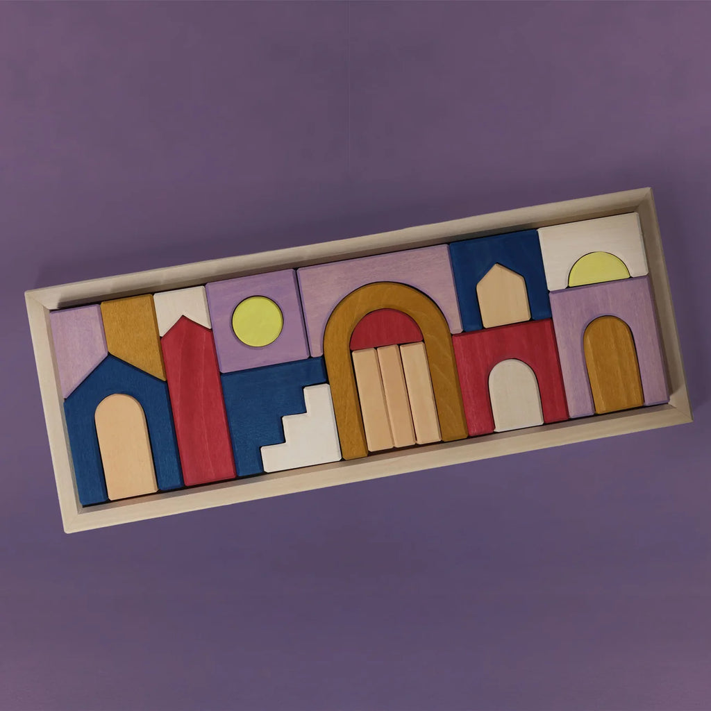 The Grimm's Panorama Building Set features vibrant geometric wooden blocks in blue, red, yellow, and natural tones, forming abstract houses and arches on a wooden tray—creating a lively scene against a purple background.