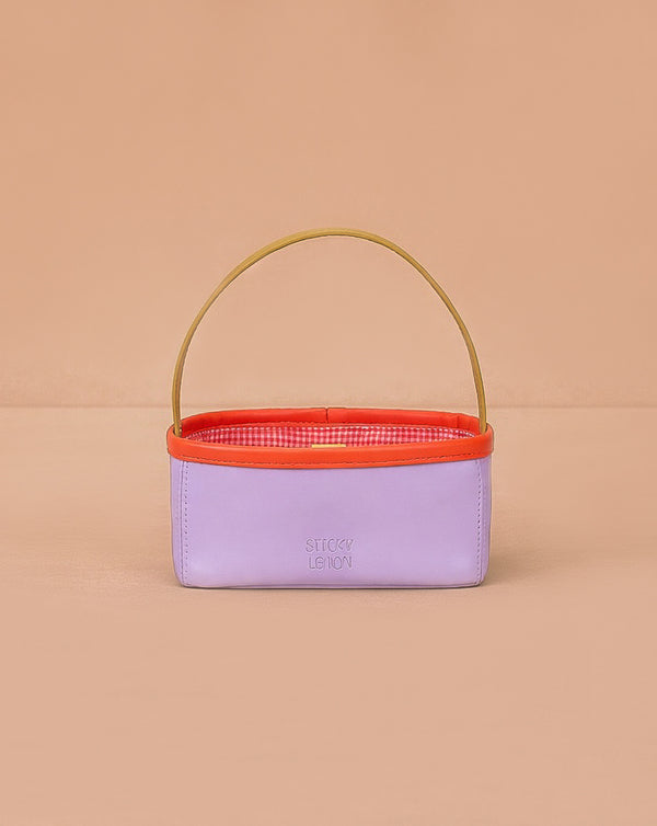The Sticky Lemon Treasure Bag - Dewy Lilac is a small rectangular bag in lavender PU leather with a yellow handle, orange rim, and red-and-white checkered interior, shown against a peach background.