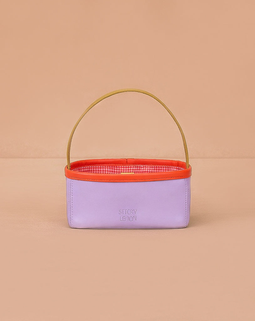 The Sticky Lemon Treasure Bag - Dewy Lilac is a small rectangular bag in lavender PU leather with a yellow handle, orange rim, and red-and-white checkered interior, shown against a peach background.