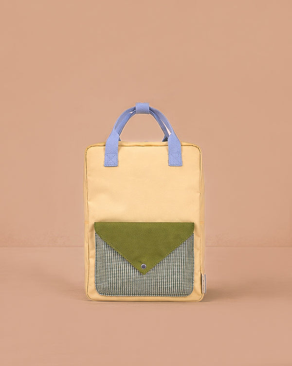 The Sticky Lemon Backpack Large - Envelope in Twiggy Beige stands upright with yellow and blue handles, a green flap pocket with a blue snap, and black-and-white checkered fabric. Made from recycled PET bags, it’s set against a beige background.