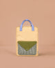 The Sticky Lemon Backpack Large - Envelope in Twiggy Beige stands upright with yellow and blue handles, a green flap pocket with a blue snap, and black-and-white checkered fabric. Made from recycled PET bags, it’s set against a beige background.