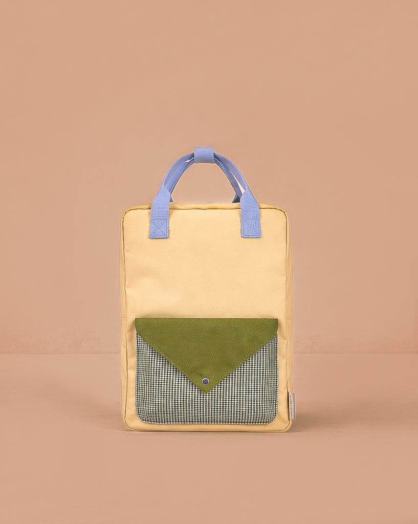 The Sticky Lemon Backpack Large - Envelope in Twiggy Beige stands upright with yellow and blue handles, a green flap pocket with a blue snap, and black-and-white checkered fabric. Made from recycled PET bags, it’s set against a beige background.