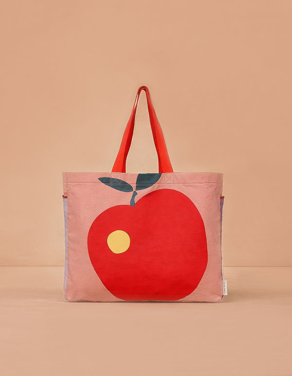 The Sticky Lemon Shopper - Apple is a colourblock tote with a large red apple, yellow dot, and green leaf on light pink. It has bold red handles and is shown against a beige background.
