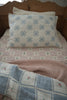 A neatly made twin bed with a large blue star pillow, a pink quilted blanket with small flowers, and the Organic Cotton Twin Size Blanket - Fleur—crafted in Portugal—folded at the foot of the bed.