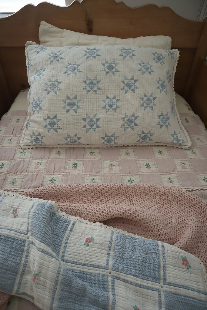 A neatly made twin bed with a large blue star pillow, a pink quilted blanket with small flowers, and the Organic Cotton Twin Size Blanket - Fleur—crafted in Portugal—folded at the foot of the bed.