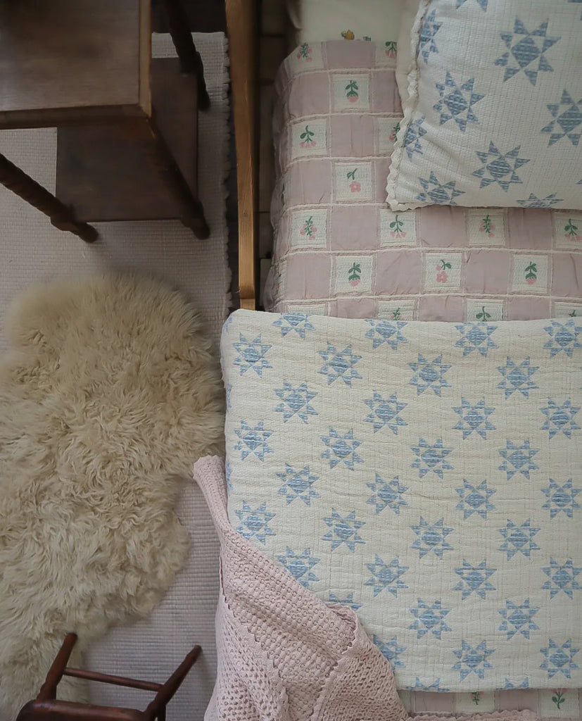 A cozy bedroom features a twin bed with the Organic Cotton Twin Size Blanket - Fleur, a patchwork pillow, a knitted pink throw, a wooden nightstand, and a fluffy cream rug on the floor.