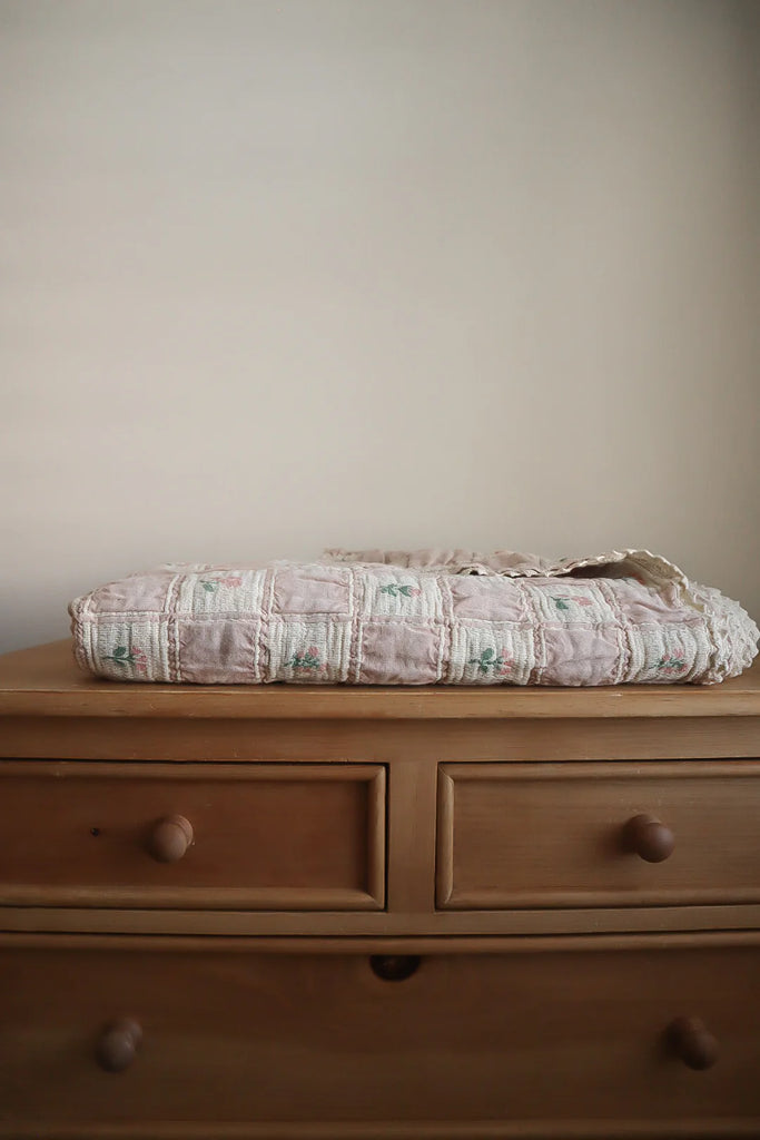 The Organic Cotton Twin Size Blanket - Fleur, featuring soft colors and a floral pattern, is neatly folded atop a light wood two-drawer dresser with round knobs, set against a plain beige wall.