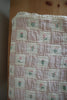 The Organic Cotton Twin Size Blanket - Fleur, made in Portugal, features a pink and cream checkered pattern with embroidered flowers in each square and lace trim, shown draped on a wooden surface.