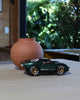 A Car Mini KEN ROGERS Metallic Green 7in collectible sits on a light wooden table beside a round, matte terracotta vase. Green foliage and sunlight brighten the background.