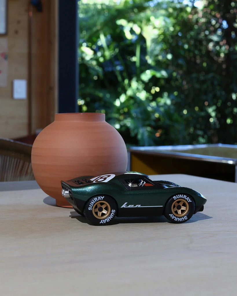 A Car Mini KEN ROGERS Metallic Green 7in collectible sits on a light wooden table beside a round, matte terracotta vase. Green foliage and sunlight brighten the background.