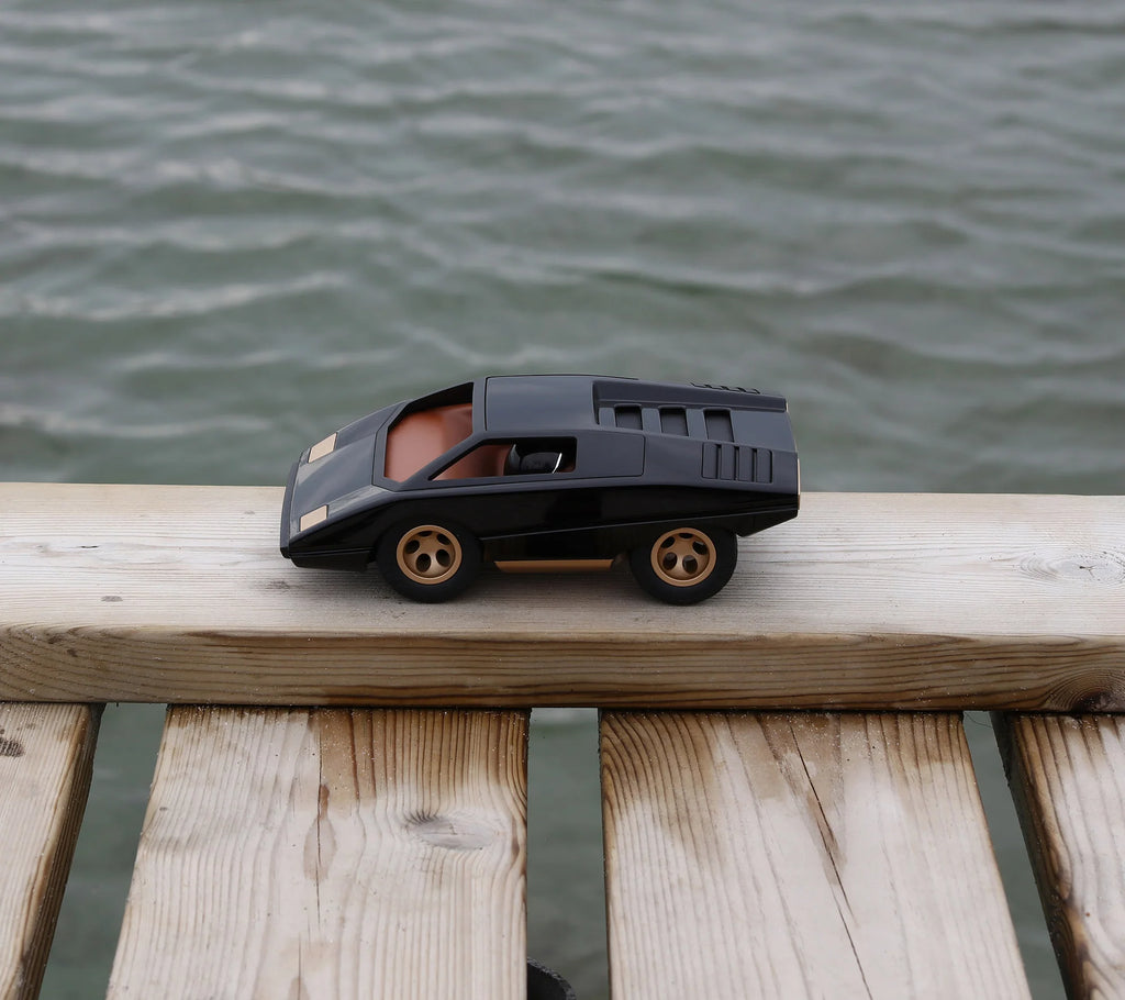 The Playforeer UFO Cannone Car in Black, featuring tan windows and wheels, sits on a wooden dock above rippling water, evoking 70’s concept car style.