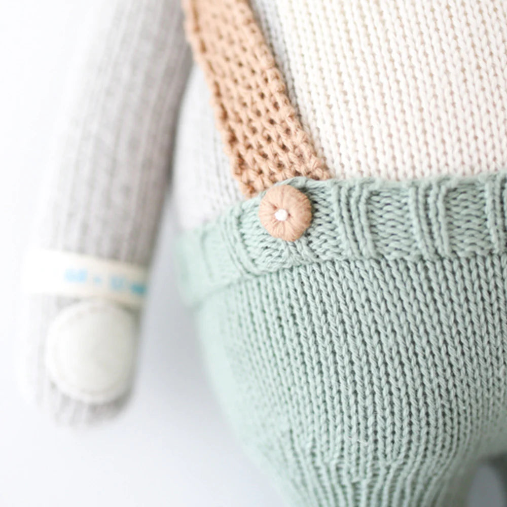 Close-up of Cuddle + Kind Quinn the Koala, hand knit in Peru, featuring mint green pants with a wooden button, tan suspenders, and a light grey arm with a white patch near the wrist. Made from 100% cotton yarn.