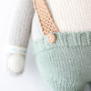 Close-up of Cuddle + Kind Quinn the Koala, hand knit in Peru, featuring mint green pants with a wooden button, tan suspenders, and a light grey arm with a white patch near the wrist. Made from 100% cotton yarn.