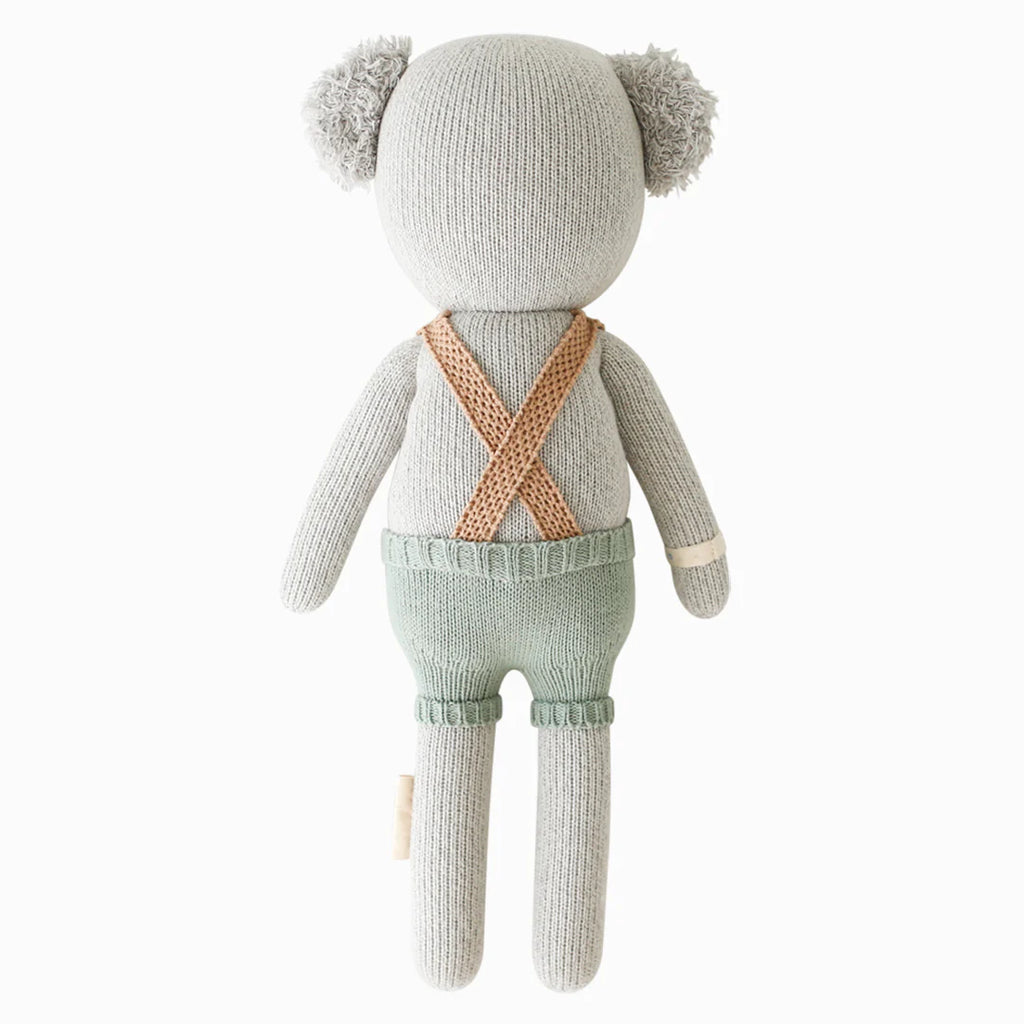 Cuddle + Kind Quinn the Koala is a hand-knit gray koala doll, shown from the back wearing green shorts with tan suspenders and a beige patch on its left arm. Handmade in Peru from 100% cotton yarn.