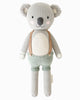 Cuddle + Kind Quinn the Koala is a hand-knit doll made from 100% cotton yarn, featuring grey fur, round ears, pink cheeks, a white belly, and dressed in green shorts with tan suspenders.