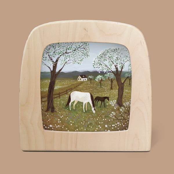 A pastoral scene depicted in the "Silhouette Only - Spring Frolic Silhouette for Toverlux Lamp" features enchanting silhouettes of a white horse and foal grazing in a flower-filled meadow, surrounded by blossoming trees, with a small house visible under a clear sky.