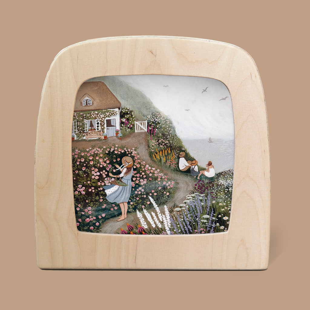 The product, "Silhouette Only - Rose Cottage Silhouette for Toverlux Lamp," depicts a seaside cottage nestled among lush gardens. In the foreground, a woman with a basket of flowers stands while two people admire the ocean and sailboat, with birds soaring in the sky above this enchanting scene crafted in translucent material.