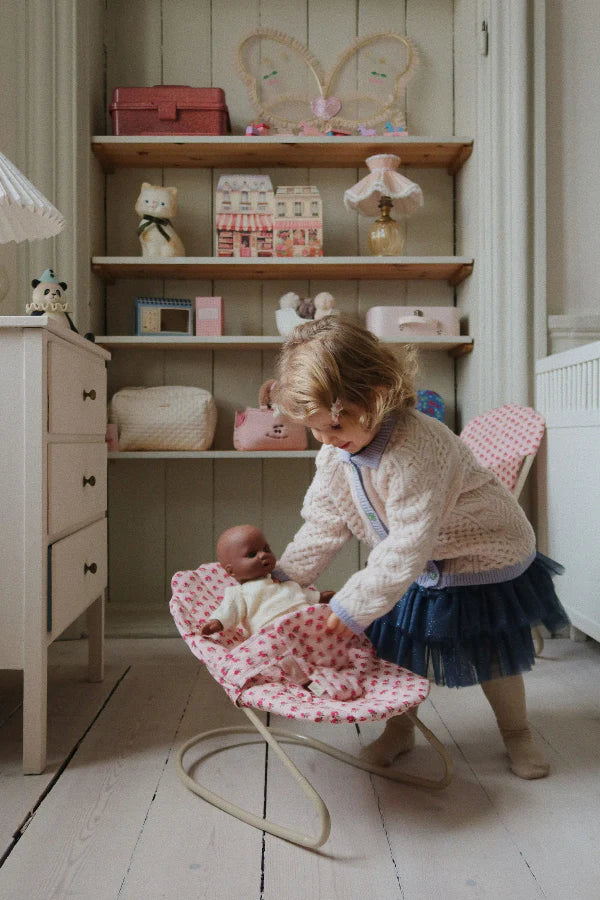 A young girl in a blue skirt and white sweater gently places a baby doll in the Konges Sloejd Doll Bouncer - Rosie Rose, a pink polka dot bouncer, in a cozy children’s room decorated with toys and shelves.