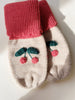 A pair of Sonja Knit Mittens - Beige Melange with a cherry design, lying on a white surface illuminated by natural light. The top of the mittens flaunts a ribbed pink cuff.