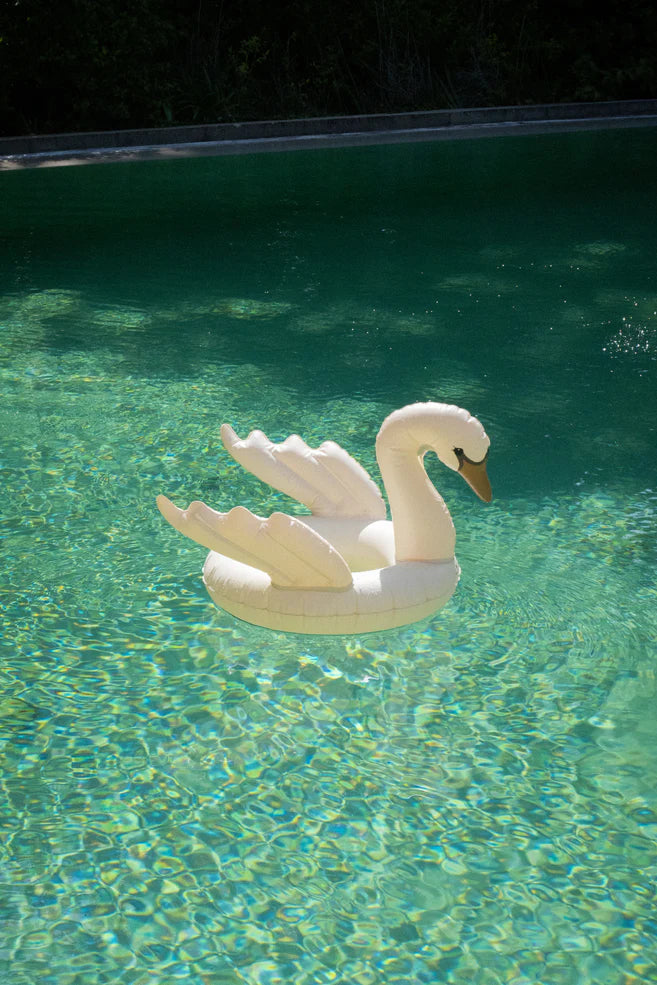 An inflatable swim ring - Swan drifts on the sparkling turquoise waters of a sunlit swimming pool, surrounded by lush greenery.