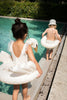 Two young children, one wearing a white Swan Inflatable Swim Ring and a hat, the other in a similar swim ring, sit on the edge of a sunlit pool, preparing to enter the water