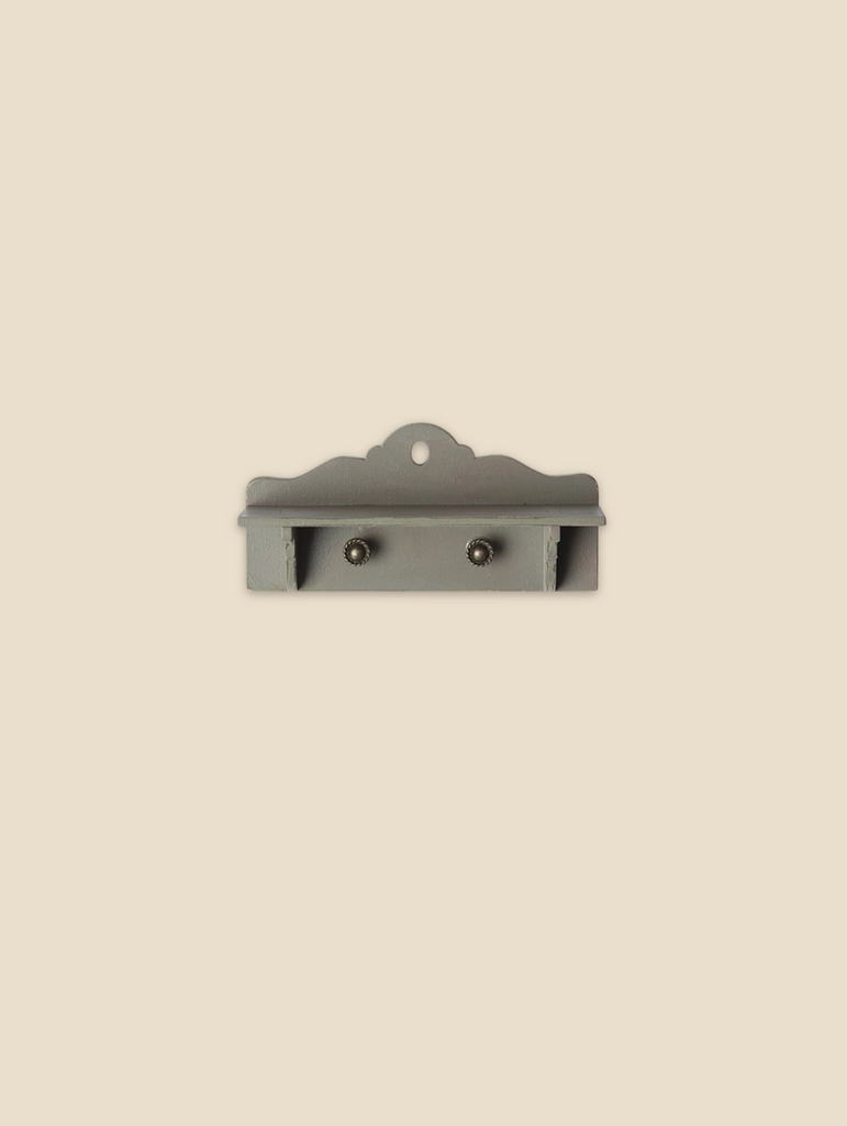 A small, decorative, wall-mounted wooden shelf with two hooks. The gray Maileg Miniature Dollhouse Shelf features a scalloped top edge and a central hole for hanging. Its minimalist design is perfect for holding small items and hanging keys or accessories, adding charm to any Maileg Dollhouse decor. Placed on a light beige background.