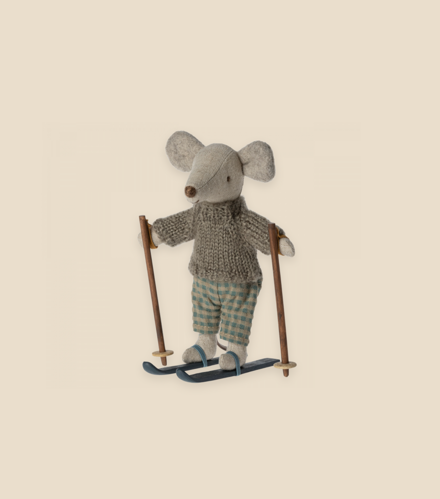 A small, plush mouse stands on skis, holding wooden poles. The Maileg Christmas Winter Mouse With Ski Set, Big Brother features the mouse dressed in a knitted gray sweater and checkered pants, all made from recycled polyester. The background is a plain, light beige color.