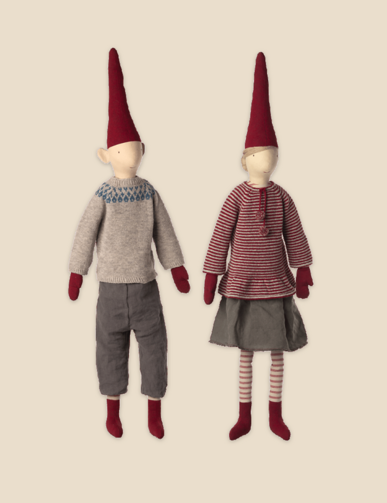 Two Maileg Christmas Mega Pixy dolls standing against a light background, each wearing a red hat. The left doll is in a grey sweater and pants made from exclusive materials, and the right doll wears a striped dress.