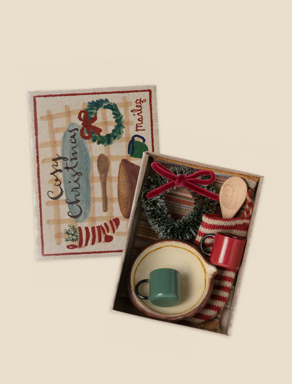 Introducing the Maileg Cosy Christmas Set: a festive gift box that includes charming Christmas accessories such as a small green mug, red mug, wooden spoon, plate, and a knitted wreath adorned with a red bow. The lid is beautifully decorated with holiday ornaments and features "Merry Christmas" in cursive, evoking the warmth of a cozy gingerbread house scene.