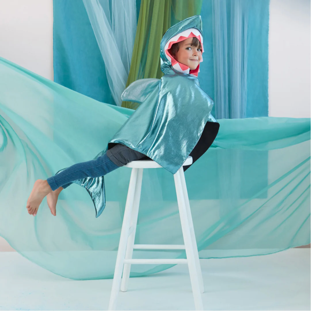 A child dressed in the Meri Meri Shark Costume - Final Sale, complete with felt teeth detail, sits on a white stool with legs playfully dangling, surrounded by draped blue and green fabric that suggests water.