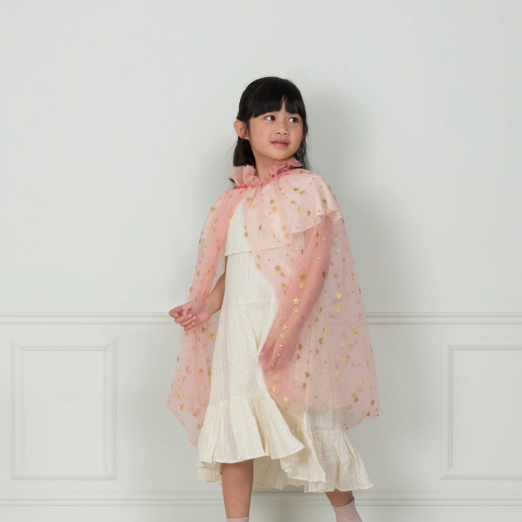 A young girl in a Meri Meri Pink Tulle Star Cape Costume stands in front of a white paneled wall, looking to the side with a gentle smile.